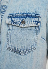 Κοντινό πλάνο ανοιχτού γαλάζιου πουκαμίσου από denim με μία τσέπη με καπάκι και μαύρο μεταλλικό κουμπί, εμφανείς ραφές και ελαφρώς ξεθωριασμένη υφή.