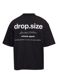 T-shirt noir oversize avec texte blanc dans le dos : "drop.size," « Édition limitée », « vêtements authentiques », et « Opération mondiale © Fondé en 2020 ».