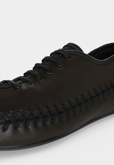 Chaussure en cuir noir avec lacets noirs et détail de surpiqûre visible le long du côté, présentée sur un fond uni.