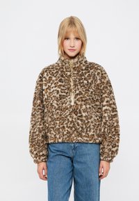 Brązowy polarowy sweter z jasnobrązowymi i ciemnobrązowymi plamami, wysoki kołnierz, pół-zamek i szerokie rękawy, zestawiony z niebieskimi dżinsami.