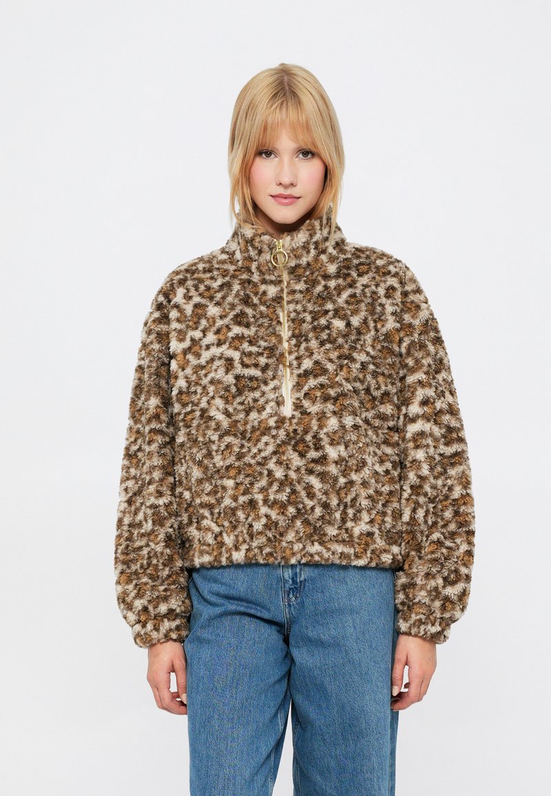 Brązowy polarowy sweter z jasnobrązowymi i ciemnobrązowymi plamami, wysoki kołnierz, pół-zamek i szerokie rękawy, zestawiony z niebieskimi dżinsami.