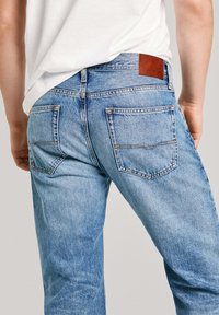 Svetlo modri zahodni jeans s sproščenim krojem, ki imajo zadnje žepe in rjav usnjen žep na pasu. Vidna obrabljena tekstura.