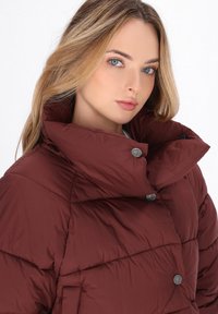 Giacca trapuntata bordeaux con colletto alto, caratterizzata da bottoni a pressione color argento, texture trapuntata e design leggero.