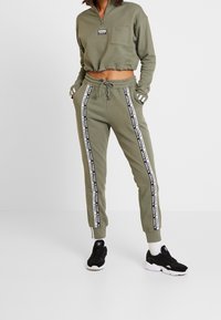 Olivgrön kort sweatshirt med framficka och halvdragkedja, kombinerad med matchande joggers med vita sidostriper och dragsko i midjan.