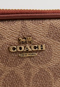 Portefeuille en cuir marron avec surface texturée, logo doré représentant une calèche tirée par des chevaux avec l'inscription "COACH NEW YORK" en dessous, et fermeture éclair rouge contrastante.