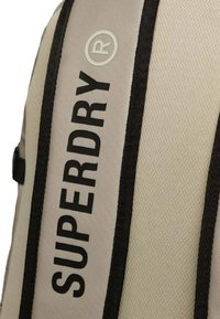 Zaino rettangolare beige con spallacci neri con il logo "SUPERDRY". Il materiale appare testurizzato, progettato per la durata.
