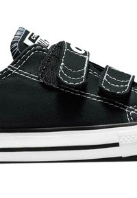 Zapatillas de lona negras con dos tiras de Velcro, costuras blancas en contraste y una suela de goma blanca que presenta dos rayas negras.