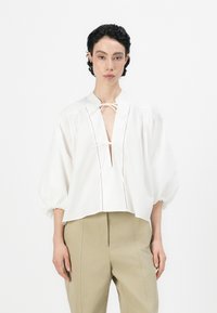 Camicetta bianca con una vestibilità ampia, maniche a sbuffo e scollatura a V profonda. Presenta una chiusura a fiocco e righe verticali sul fronte.