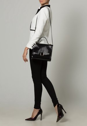 Donna che indossa un blazer bianco, pantaloni neri e tacchi alti, mentre porta una borsa nera lucida con manico e tracolla.