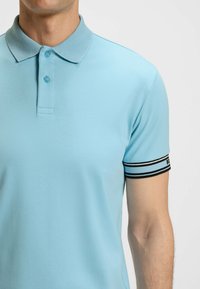 Helles blaues Poloshirt aus glattem Stoff, mit strukturiertem Kragen und schwarz-weißen Streifenakzenten an den Ärmeln. Mit zwei Knöpfen.