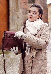 Burgundská křížová taška vyrobená z syntetického materiálu se zipem nahoře, držena v rukavici vedle prošívaného béžového kabátu.