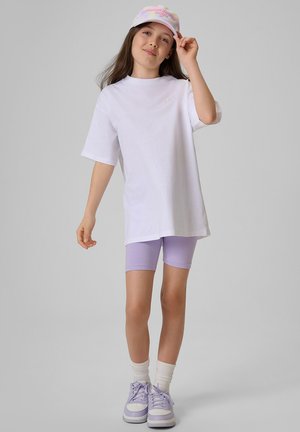 Dziewczyna ubrana w białą, oversize koszulkę, fioletowe krótkie spodenki kolarskie, pasujące tenisówki, białe skarpetki i pastelową czapkę tie-dye, poprawiająca czapkę jedną ręką.