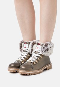 Botines metálicos de bronce con cordones blancos, puño de piel sintética, acentos de hardware dorado y suela de goma texturizada con profundo relieve.
