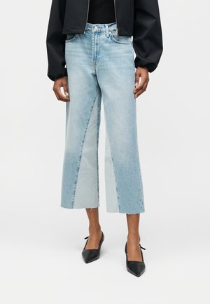 Személy, aki világoskék, széles szárú, rövidített farmernadrágot visel kéttónusú farmer panelekkel és fekete, hegyes orrú magas sarkú cipőket, egyszerű háttér előtt áll.