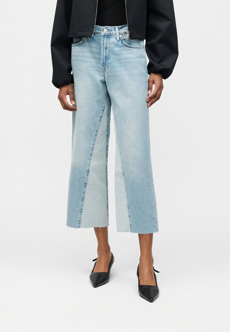 Άτομο που φοράει φαρδιά cropped τζιν σε ανοιχτό μπλε με δίχρωμα τζιν πάνελ και μαύρα μυτερά τακούνια, στέκεται μπροστά σε απλό φόντο.