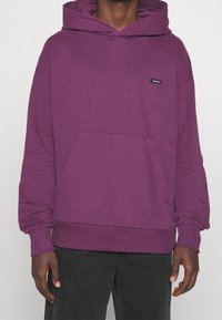 Paarse hoodie met een kangoeroezak, ribgebreide manchetten en zoom, en een klein merkje op de borst. Gemaakt van een zachte stof met veel katoen.