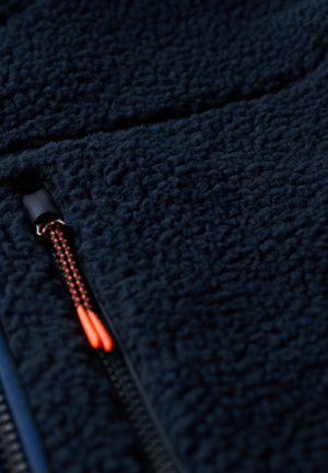 Tissu polaire bleu marine avec une surface texturée. Présente une fermeture éclair noire avec une tirette orange. Le design est lisse et doux.