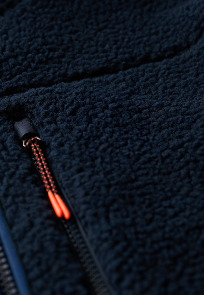 Marine fleece stof met een gestructureerd oppervlak. Voorzien van een zwarte rits met een oranje trekband. Het ontwerp is glad en zacht.