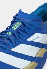 Blauer Sportschuh mit texturierter Oberseite, weißen Streifen, grünem Markenlogo und goldener Sohle mit verstärkten Nähten.