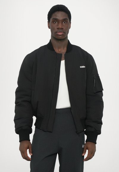 Schwarze Bomberjacke mit einem Reißverschluss vorne, gerippten Bündchen und einer Brusttasche. Hergestellt aus glattem Stoff, mit Logodesign auf der Brust.