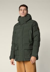 Uomo che indossa una giacca invernale imbottita verde scuro con colletto alto, tasche frontali e pantaloni beige, in piedi davanti a uno sfondo semplice.