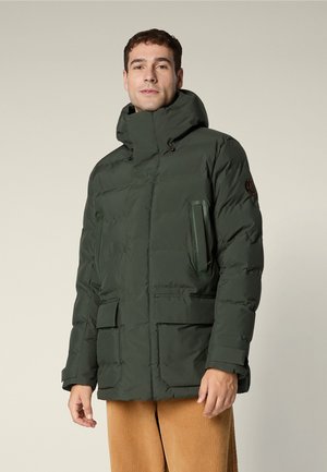 Man in een donkergroene gewatteerde winterjas met hoge kraag, voorzakken en beige broek, staand tegen een effen achtergrond.
