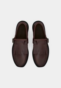 Mocassins en cuir marron avec des accents à franges et une semelle en caoutchouc noir. Dotés d'une boucle sur le côté et de détails cousus sur le dessus.