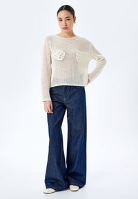 Strickpullover in Off-White mit floralen Akzenten auf der Brust, kombiniert mit hoch taillierten, weit geschnittenen blauen Jeans und cremefarbenen Schuhen.