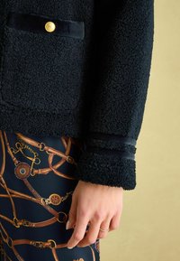 Čierna fleecová bunda s textúrovaným povrchom, s zlatým gombíkom a predným vreckom. Rukáv má sametový akcent a pruhy.