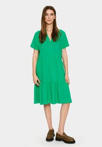 Rochie verde, până la genunchi, cu decolteu în V, mâneci scurte și fustă cu volane. Textură netedă, asortată cu pantofi săi brown.