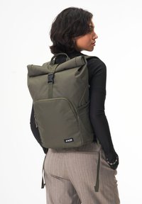 Grüner Rolltop-Rucksack aus strapazierfähigem Material, mit Seitentaschen aus Mesh, verstellbaren Trägern und einer Fronttasche mit Reißverschluss. Logo auf der Vorderseite.