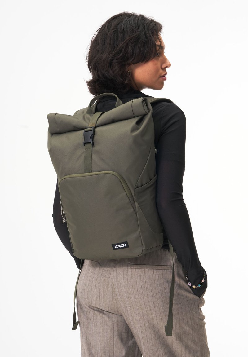 Grüner Rolltop-Rucksack aus strapazierfähigem Material, mit Seitentaschen aus Mesh, verstellbaren Trägern und einer Fronttasche mit Reißverschluss. Logo auf der Vorderseite.