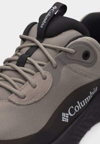 Ανοιχτό καφέ αθλητικό παπούτσι Columbia με επάνω μέρος από δίχτυ, επενδεδυμένη μαύρη γλώσσα, στρογγυλά κορδόνια και μαύρη υφασμένη σόλα, εμφανιζόμενο σε κοντινή γωνία.