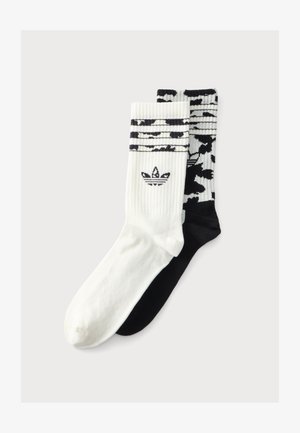 Paire de chaussettes montantes, une blanche avec des rayures noires et le logo Adidas, une noire avec un motif abstrait blanc et une zone noire au niveau des orteils.