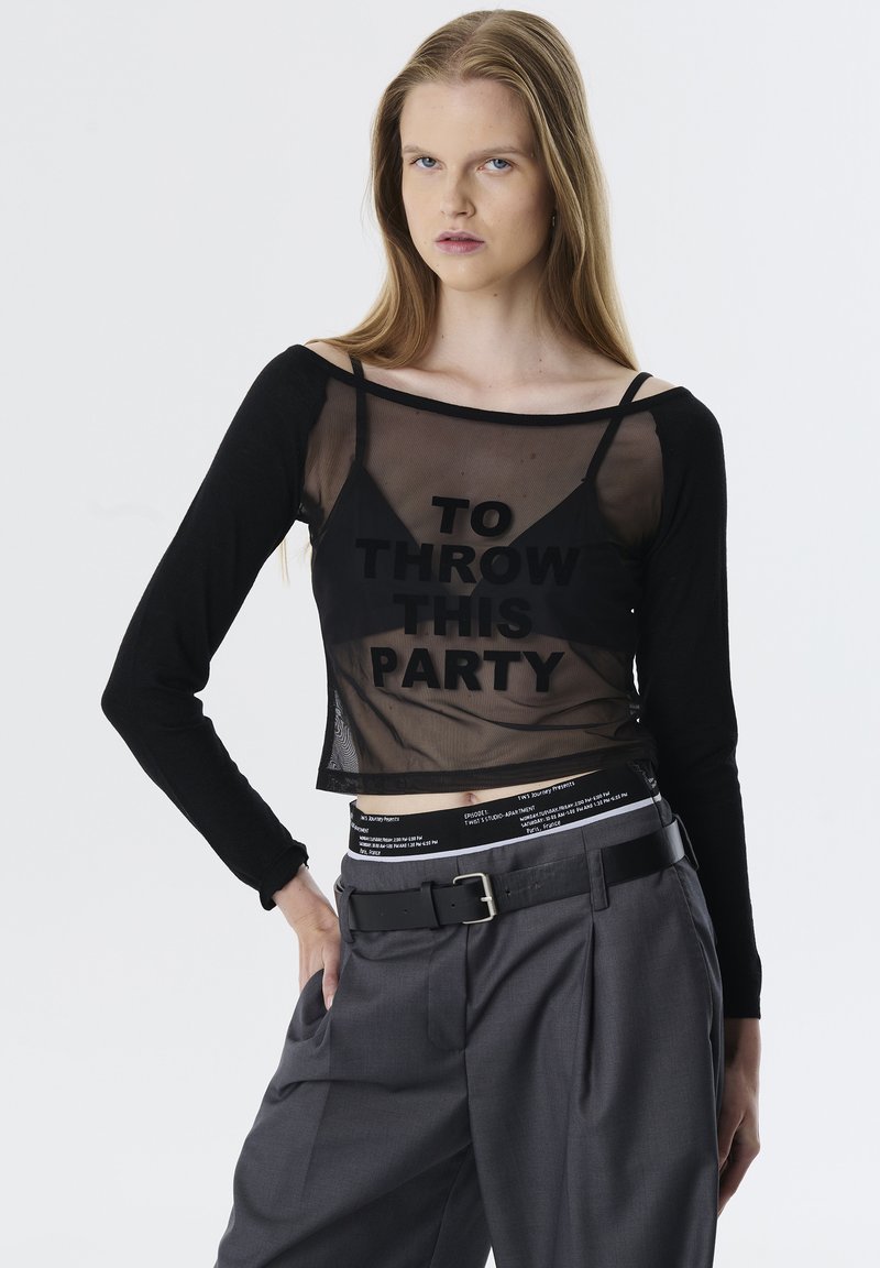 Zwarte net crop top met lange mouwen, met grote tekst "DIT FEEST GOOIEN." Gecombineerd met hoge grijze broeken en een zwarte riem.