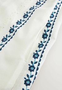 Tissu blanc avec des motifs brodés de vigne florale bleu foncé le long des bords et un subtil détail texturé à proximité.
