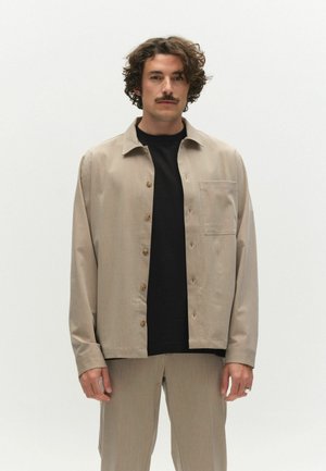 Mann mit lockigem Haar und Schnurrbart, der ein beigefarbenes Hemd über einem schwarzen T-Shirt und passende beige Hosen trägt, vor weißem Hintergrund stehend.