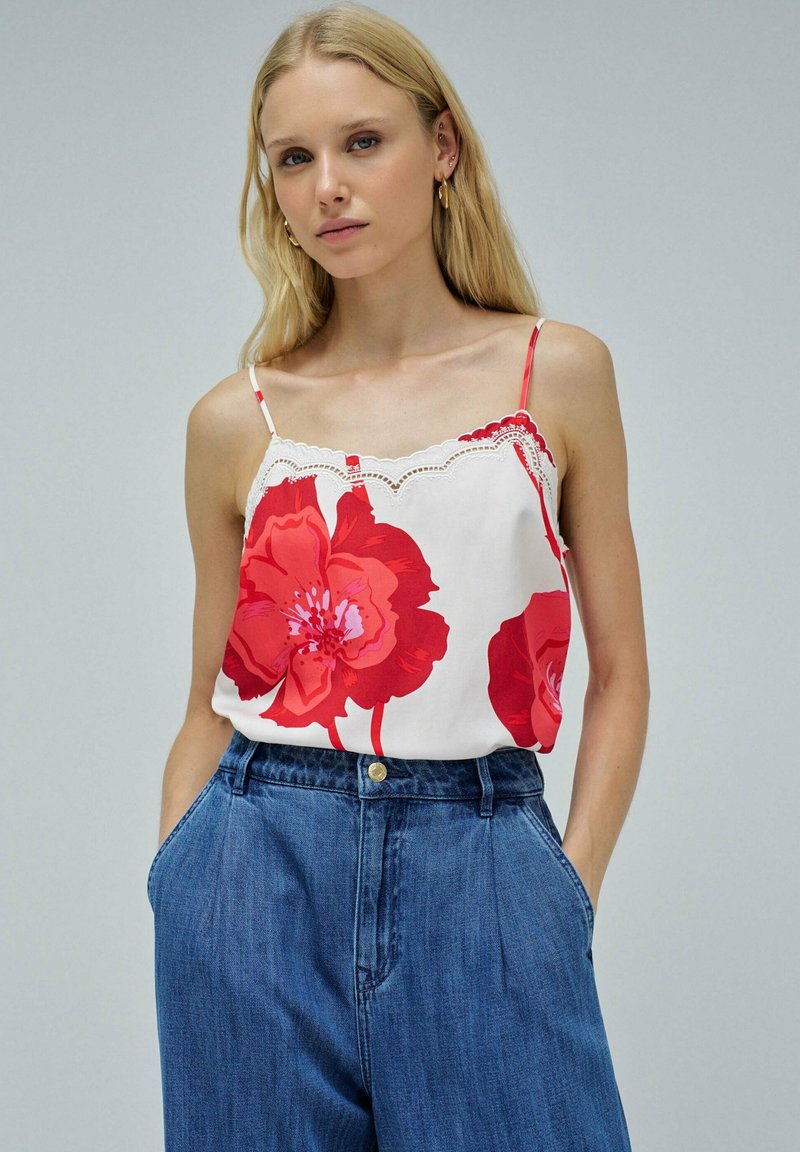 Joven mujer con cabello largo y rubio, vestida con una camiseta sin mangas blanca con grandes estampados de flores rojas y jeans de tiro alto azul, con las manos en los bolsillos.