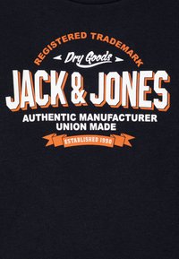 La camiseta de algodón azul marino presenta un texto en blanco y naranja en negrita; "JACK & JONES" es destacado, junto con detalles sobre la fabricación y el año de establecimiento.