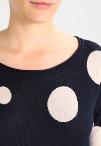 Navy sweater with large beige polka dots, featuring a round neckline and ribbed trim. Material appears knitted with a smooth texture.