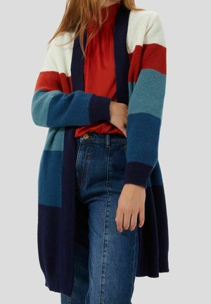 Kvinde iført lang strikket cardigan med brede, horisontale striber i hvid, rød, blå og marineblå over rød bluse og blå jeans.