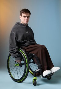 Pelēka pārāk liela kapuce, brūni bikses un baltas kedas. Persona sēž ratiņkrēslā ar melnu rāmi un zaļiem akcentiem. Gluda fona gradients.