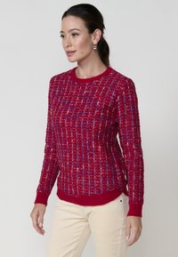 Pull en tricot rouge avec un motif texturé, présentant des accents bleus et blancs, un col et des poignets côtelés, et un ourlet légèrement incurvé.