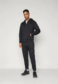 Črn atletski tracksuit iz mehkega blaga, ki vključuje zip-up kapuco in prilegajoče hlače. Obe izdelki imata subtilen logotip.