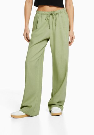 Woman wearing light green wide-leg pants with drawstring waist and white sneakers, standing against white background.