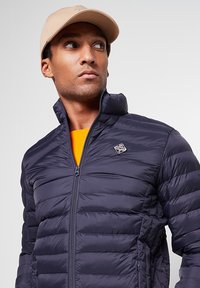 Doudoune bleu marine avec un design matelassé horizontal, fermeture éclair, col montant et un petit écusson logo. Portée par-dessus une chemise orange.