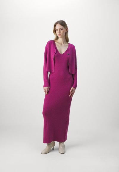 Robe en tricot fuchsia avec un décolleté en V profond et un cardigan court assorti avec des boutons. Le mannequin porte des bottines à talons beiges.