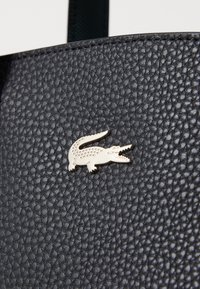 Matériau en cuir noir texturé avec un accent de logo crocodile doré. La surface présente une finition granuleuse, mettant en valeur un design distinct.