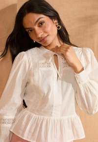 Blouse blanche avec un ourlet en peplum, motif à rayures verticales, accents en dentelle, col rond et manches longues bouffantes. Les détails incluent des points texturés.