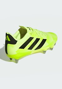 Crampons de soccer jaunes vifs avec des accents noirs et trois rayures, au design élancé, avec une tige texturée et des crampons métalliques pour une meilleure traction.